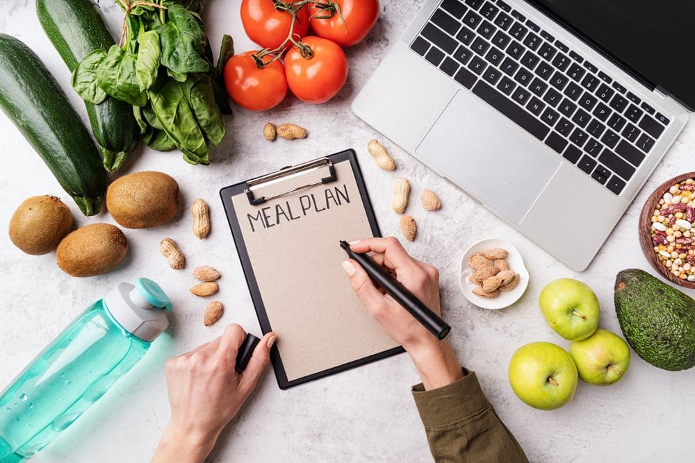 Weight lose and dieting concept. Top view of woman workspace with healthy food. Female hands writing in the notepad words meal plan top view flat lay