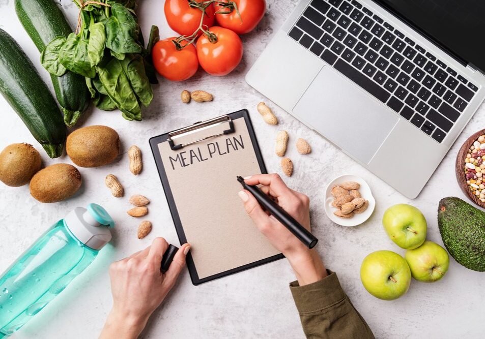 Weight lose and dieting concept. Top view of woman workspace with healthy food. Female hands writing in the notepad words meal plan top view flat lay