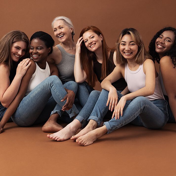 Six women of different ages sitting together in studio on brown background. Multi-ethnic group of diverse females having good times.